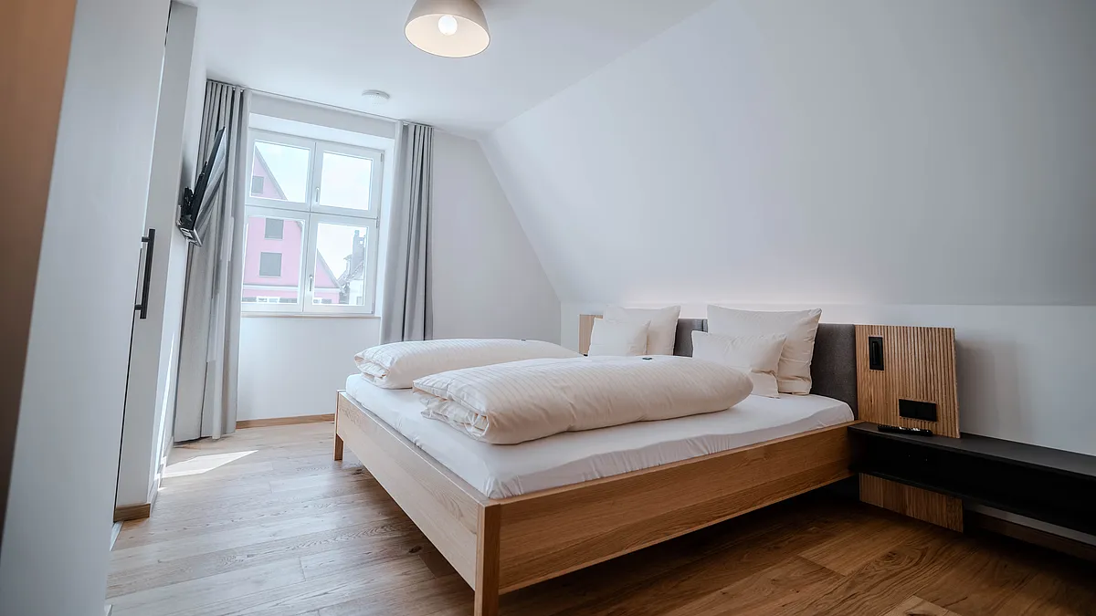 Schlafzimmer im Haus Ehemalige Sparkasse mit Doppelbett unter der Dachschräge, Pendelleuchte und Fenster mit grauen Vorhängen