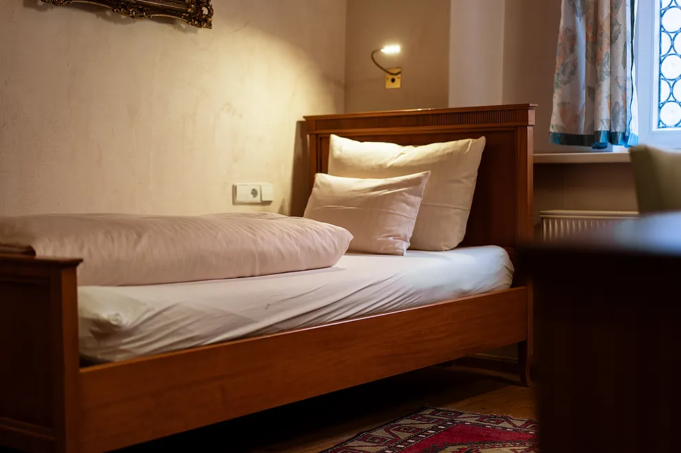 Einzelbett aus Holz mit weißer Bettwäsche und Wandleselampe im Standard-Einzelzimmer des Stammhauses