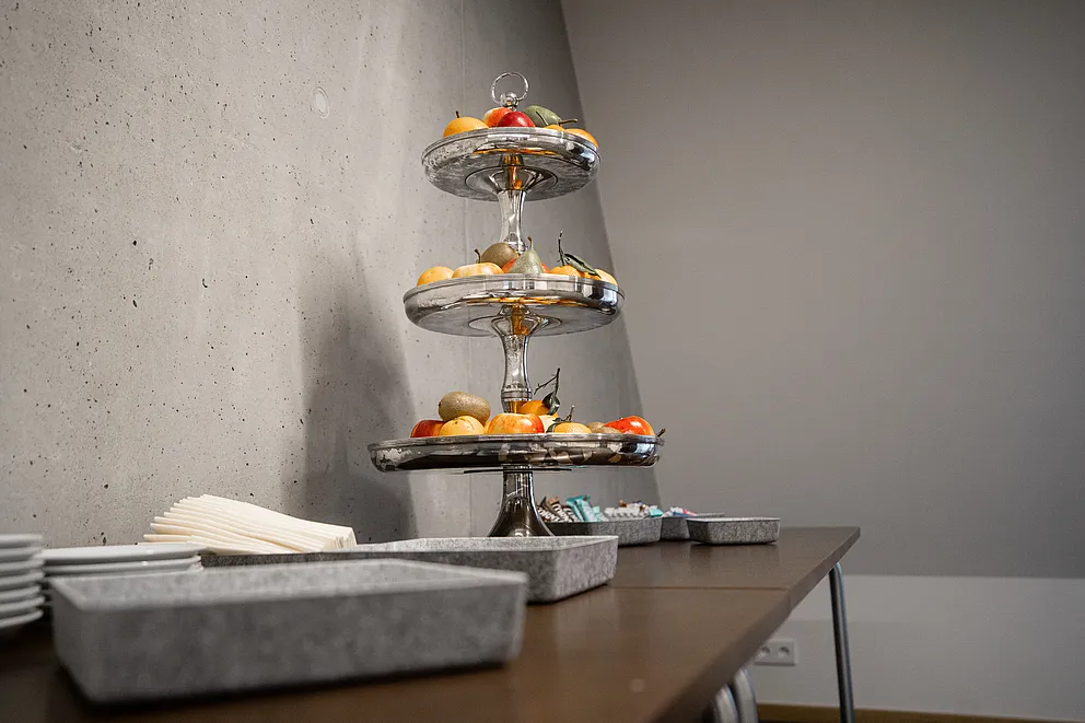 Dreistöckige silberne Etagere mit frischem Obst auf einem Holztisch mit Tellern und Servietten vor einer Betonwand