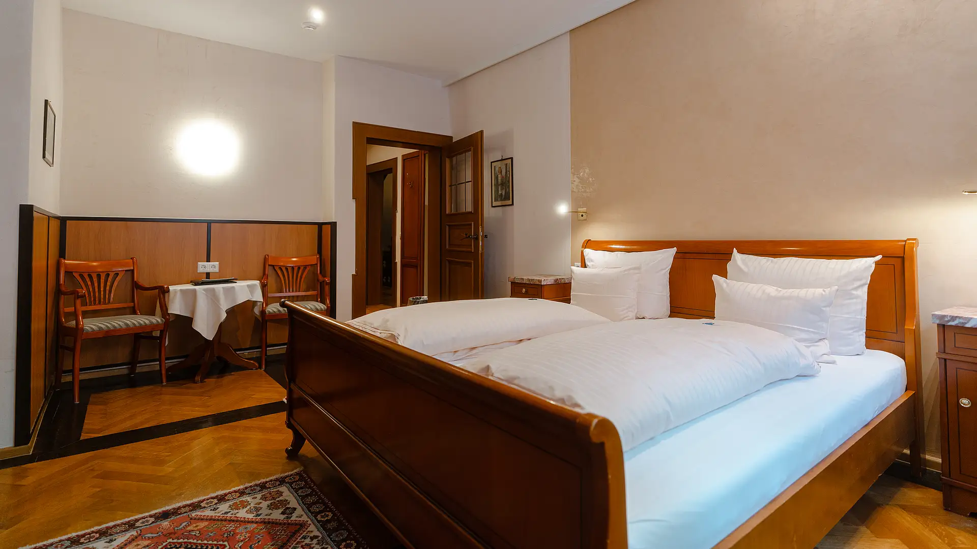 Zimmer mit Doppelbett aus rötlichem Holz, Fischgrätparkett und Sitzecke mit zwei Stühlen in holzvertäfelter Nische