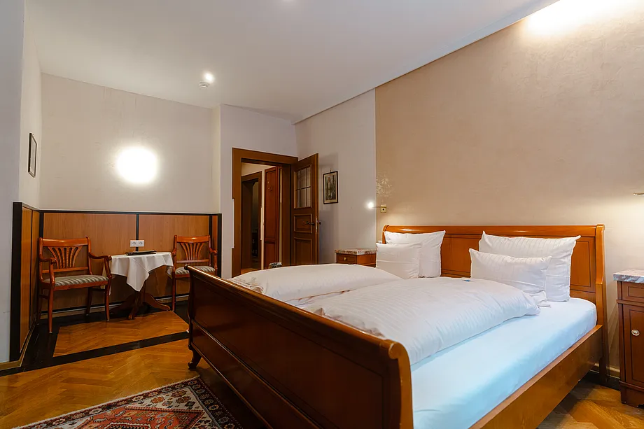 Zimmer mit Doppelbett aus rötlichem Holz, Fischgrätparkett und Sitzecke mit zwei Stühlen in holzvertäfelter Nische