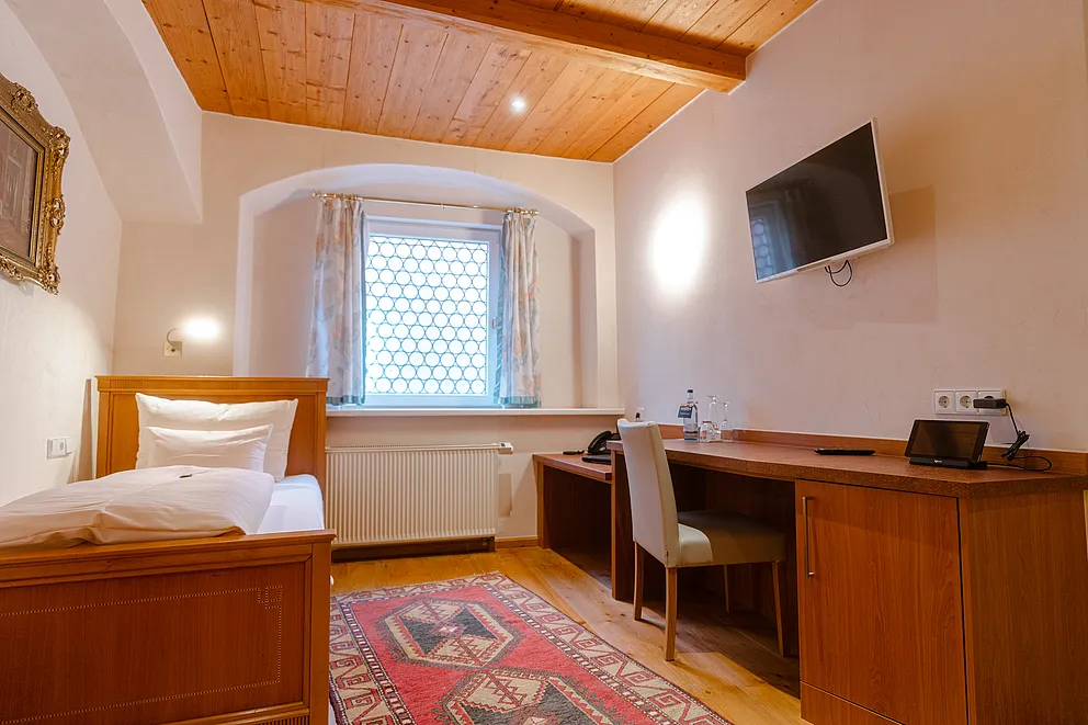 Standard-Einzelzimmer im Stammhaus mit Holzbett, Holzdecke, Schreibtisch mit Stuhl, Wandfernseher und rotem Teppich