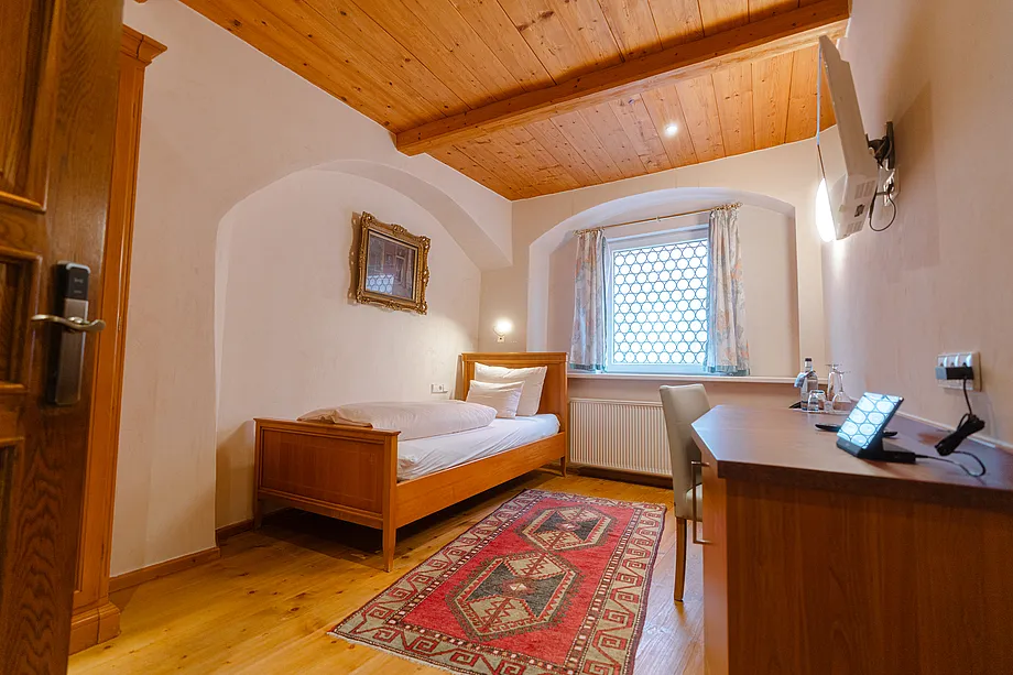 Zimmer mit Einzelbett, Holzdecke, Fenster in Rundbogennische, rotem Teppich und Schreibtisch mit Fernseher