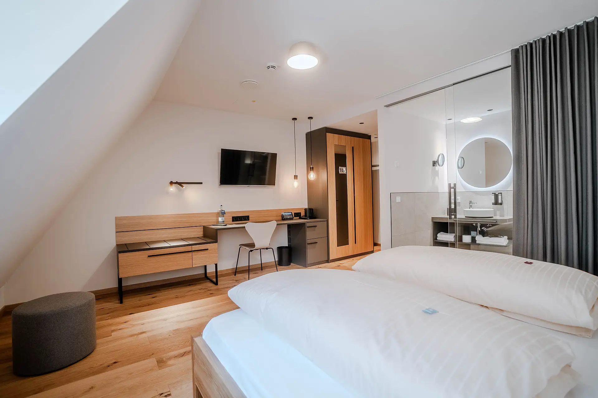 Zimmer unter Dachschräge mit Doppelbett, grauem Hocker, Schreibtisch, Wandfernseher und Bad hinter Glaswand