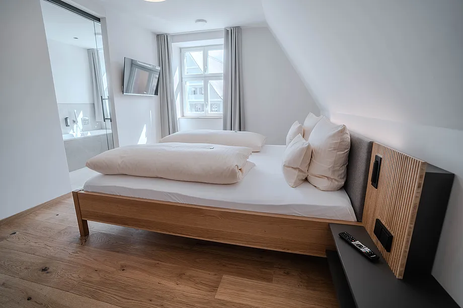 Zimmer mit Holzdoppelbett unter Dachschräge, Wandfernseher und Bad mit Badewanne hinter Glaswand