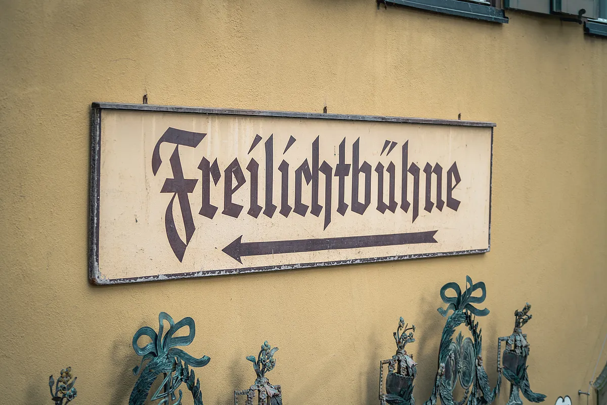 Wegweiser mit der Aufschrift Freilichtbühne in Frakturschrift an gelber Hauswand, darunter schmiedeeiserne Zierteile