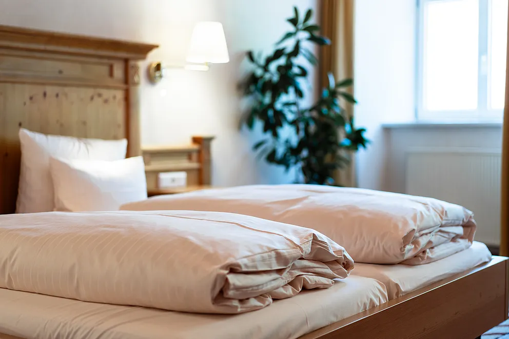 Doppelbett mit gefalteten Bettdecken und Kopfteil aus hellem Holz im Superior-Zimmer, dahinter Zimmerpflanze am Fenster