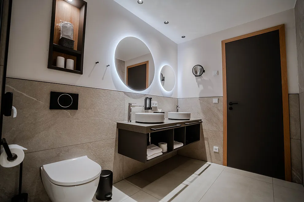 Badezimmer mit Wand-WC, schwebendem Waschtisch mit zwei Aufsatzbecken und runden Leuchtspiegeln