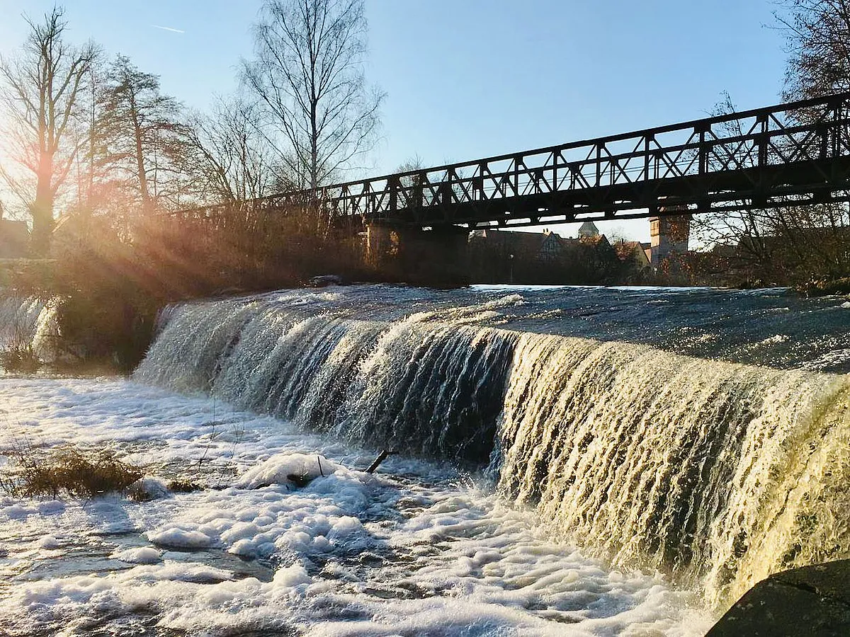 Flusswehr mit schäumendem Wasser, dahinter eine eiserne Brücke im Gegenlicht der tief stehenden Sonne