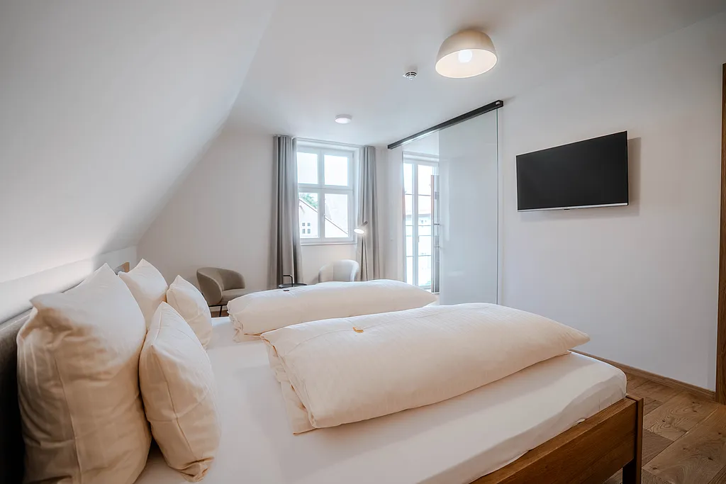 Doppelzimmer mit Holzbett unter der Dachschräge, zwei Sesseln am Fenster und Wand-TV