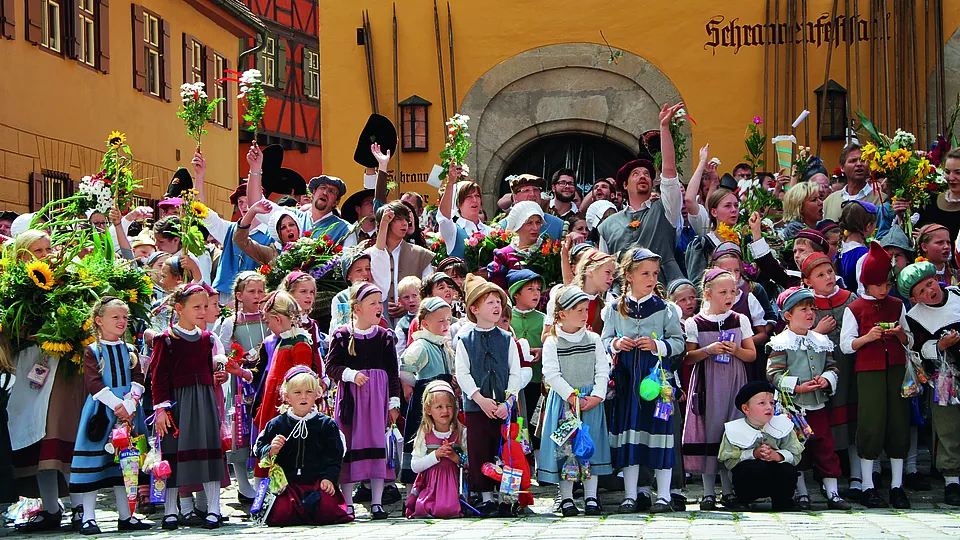 Kinder und Erwachsene in historischen Gewändern jubeln mit erhobenen Armen und Blumensträußen vor dem Schrannenfestsaal