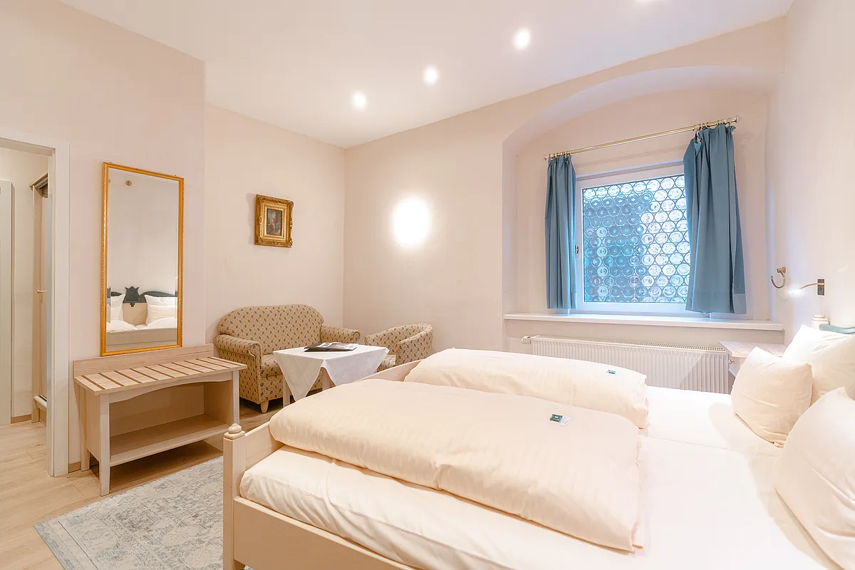 Zimmer mit hellem Holzdoppelbett, Sitzecke mit zwei Polstersesseln, Spiegel mit Goldrahmen und Fenster mit blauen Vorhängen