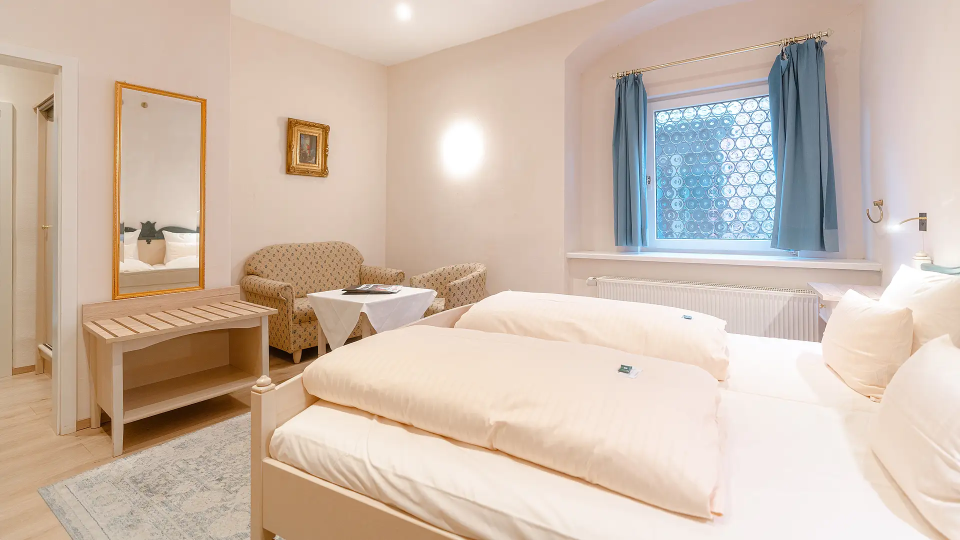 Zimmer mit hellem Holzdoppelbett, Sitzecke mit zwei Polstersesseln, Spiegel mit Goldrahmen und Fenster mit blauen Vorhängen