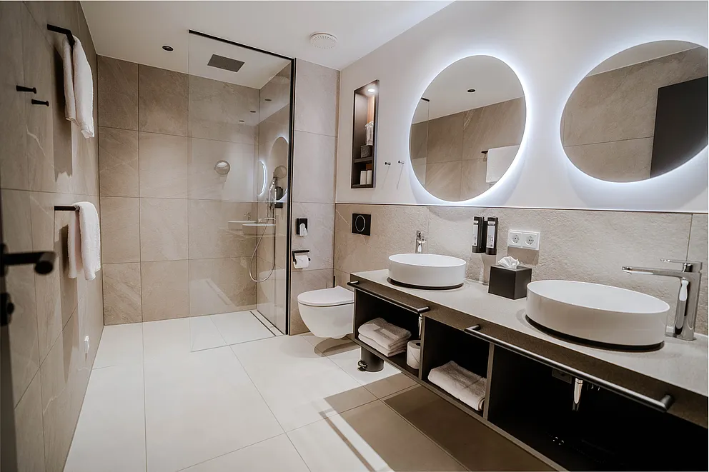 Bad der Junior-Suite im Haus Ehemalige Sparkasse mit Doppelwaschtisch, runden beleuchteten Spiegeln und Dusche mit Glaswand