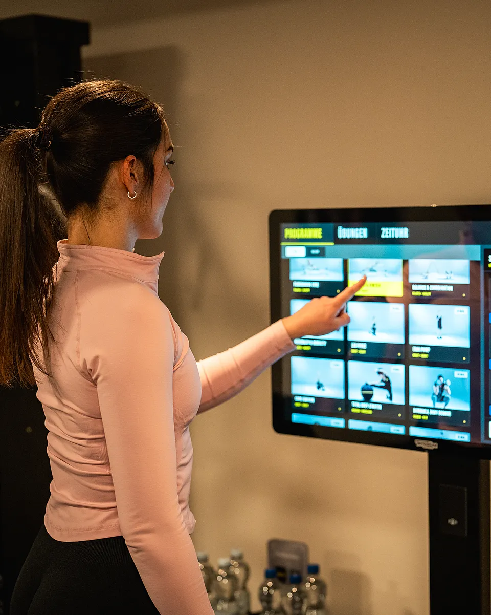 Frau wählt an einem großen Touchscreen im Fitnessraum Trainingsprogramme und Übungen aus