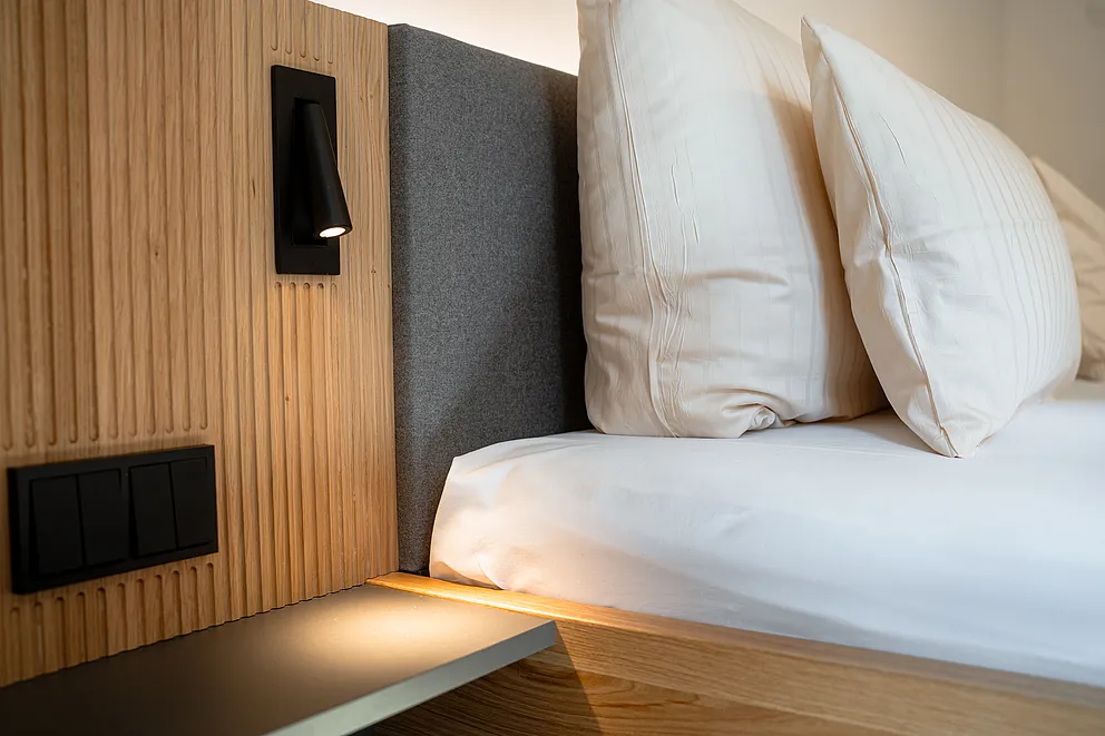Bett mit grau gepolstertem Kopfteil und weißen Kissen neben einem Holzpaneel mit schwarzer Leselampe und Schaltern
