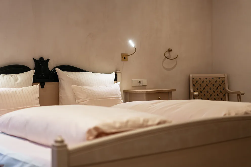 Doppelbett mit weißer Bettwäsche und dunkel verziertem Kopfteil im Standard-Doppelzimmer, daneben Nachttisch und Stuhl