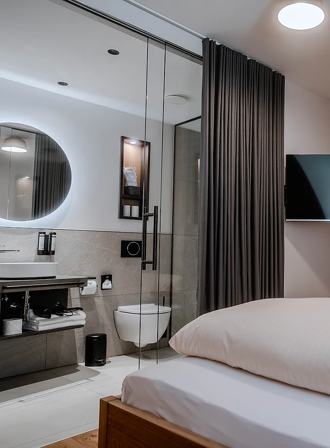 Zimmer mit Bad hinter Glaswand, rundem beleuchtetem Spiegel und Waschtisch, im Vordergrund das Bett