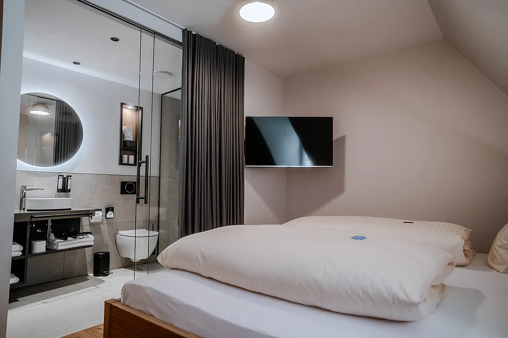 Zimmer mit Bad hinter Glaswand, rundem beleuchtetem Spiegel und Waschtisch, im Vordergrund das Bett